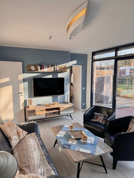 Wohnzimmer mit großer Glassfront. Ein Doppelsofa und zwei Sessel, Ein Smart-TV und ein Tisch sind dort zu finden.