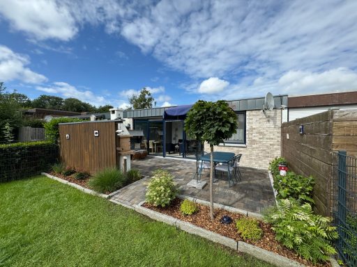 Ein grüner Streifen des Gartens im Vordergrund. Im Hintergrund Bepflanzung, ein blauer Esstisch mit 4 Stühlen und ein Strandkorb vorm Haus