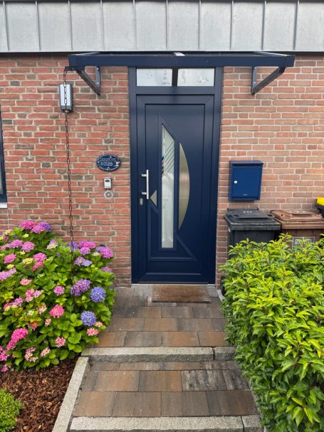 Rotes Backsteinhaus mit blauer Haustür und kleinem Vordach. Links und Rechts Bepflanzung. Rechts Neben Haustür: Briefkasten und Mülltonen