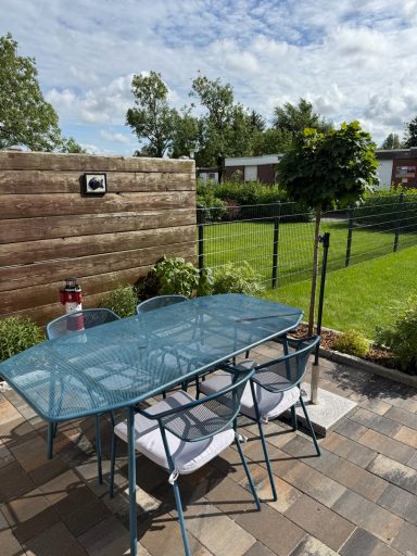 Ein blauer Garten-Esstisch aus Metall mit Vier Stühlen und Sitzauflagen. Blick in den eingezäunten Garten