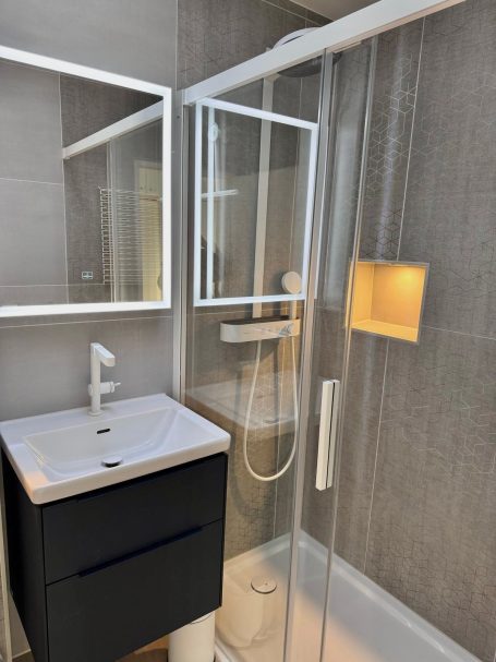 Ein Badezimmer mit Waschtisch mit 2 Schubladen, Ein großer Spiegel inklusive Beleuchtung, eine Glaswand hinter der eine Regendusche ist. 