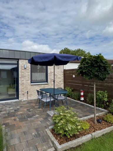 Ein blauer Garten-Esstisch aus Metall mit Vier Stühlen und Sitzauflagen unter einem blauen Sonnenschirm vor dem Wohnhaus.