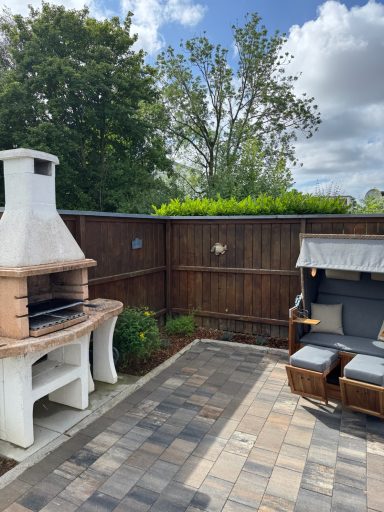 Ein Kamingrill auf einer gepflasteren Terasse. Rechts daneben steht ein Strandkorb.