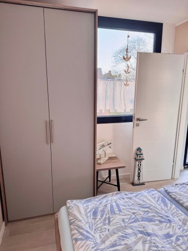 Das Fußende des Bettes ist zu sehen. Rechts vom Bett ist die Tür, links daneben ein kleiner Tisch und daneben ein großer Doppeltürer-Schrank