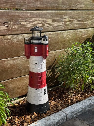 Ein Rot-Weißer Dekoleuchturm in einem Beet vor einer Holzwand.
