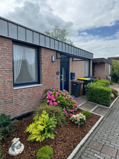 Rotes Backsteinhaus mit blauer Haustür und kleinem Vordach. Links und Rechts Bepflanzung. Rechts Neben Haustür: Briefkasten und Mülltonen