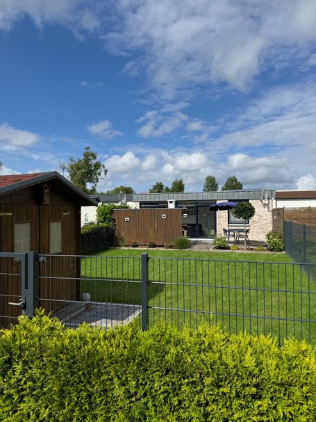 Blick in den Garten über einen Zaun und eine Hecke hinweg. Links ist ein Gartenhaus und im Hintergrund das Haus mit Terrasse.