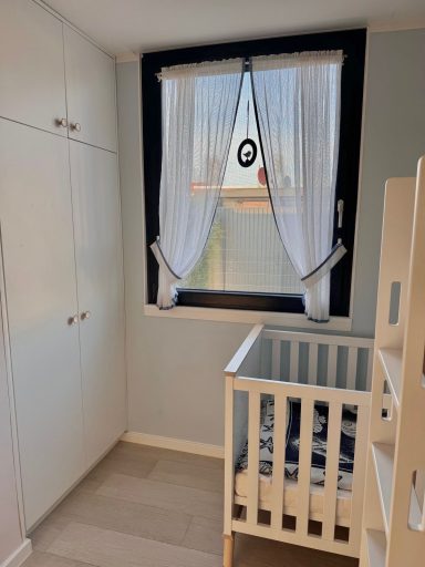 Links ist ein großer Wandschrank mit 2 Türen und darüber zwei kleinen Türen. Rechts daneben ein Fenster und weiter rechts ein Babybett